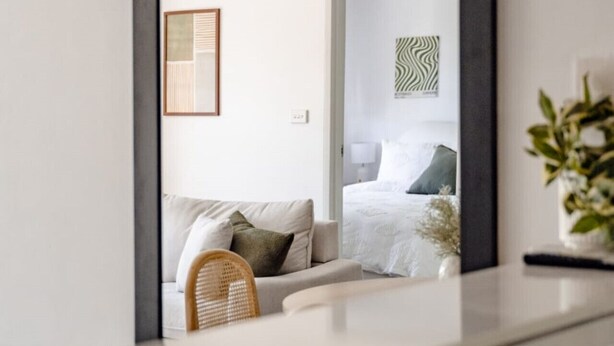 A modern interior with a large mirror reflecting a bedroom, a wicker chair, and a hanging light fixture. - Modern Neutral Haven with Premium Amenities (Griffith)