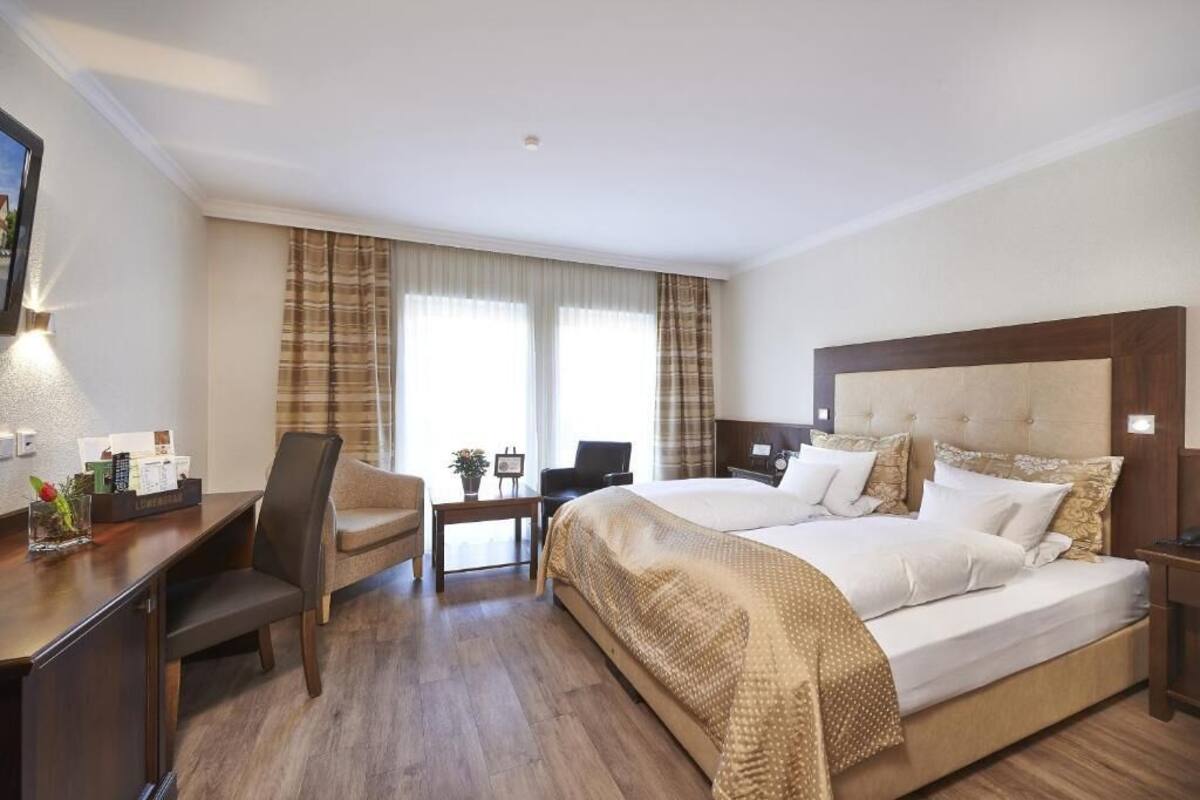 Habitación de hotel con una cama grande, un escritorio con silla, una mesita y una ventana con cortinas.
