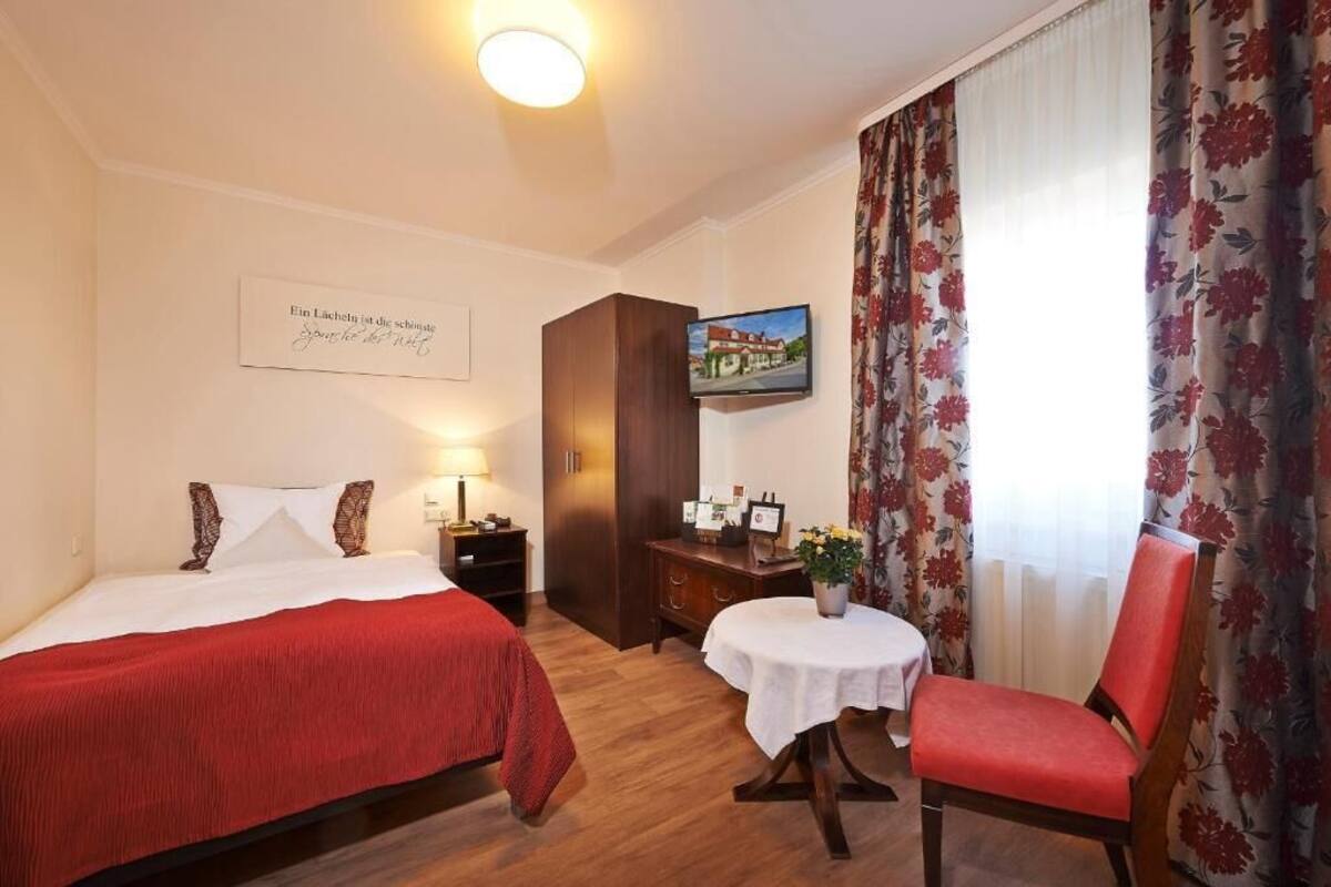 Habitación de hotel con cama, una silla, una mesita, televisión y ventana con cortinas.