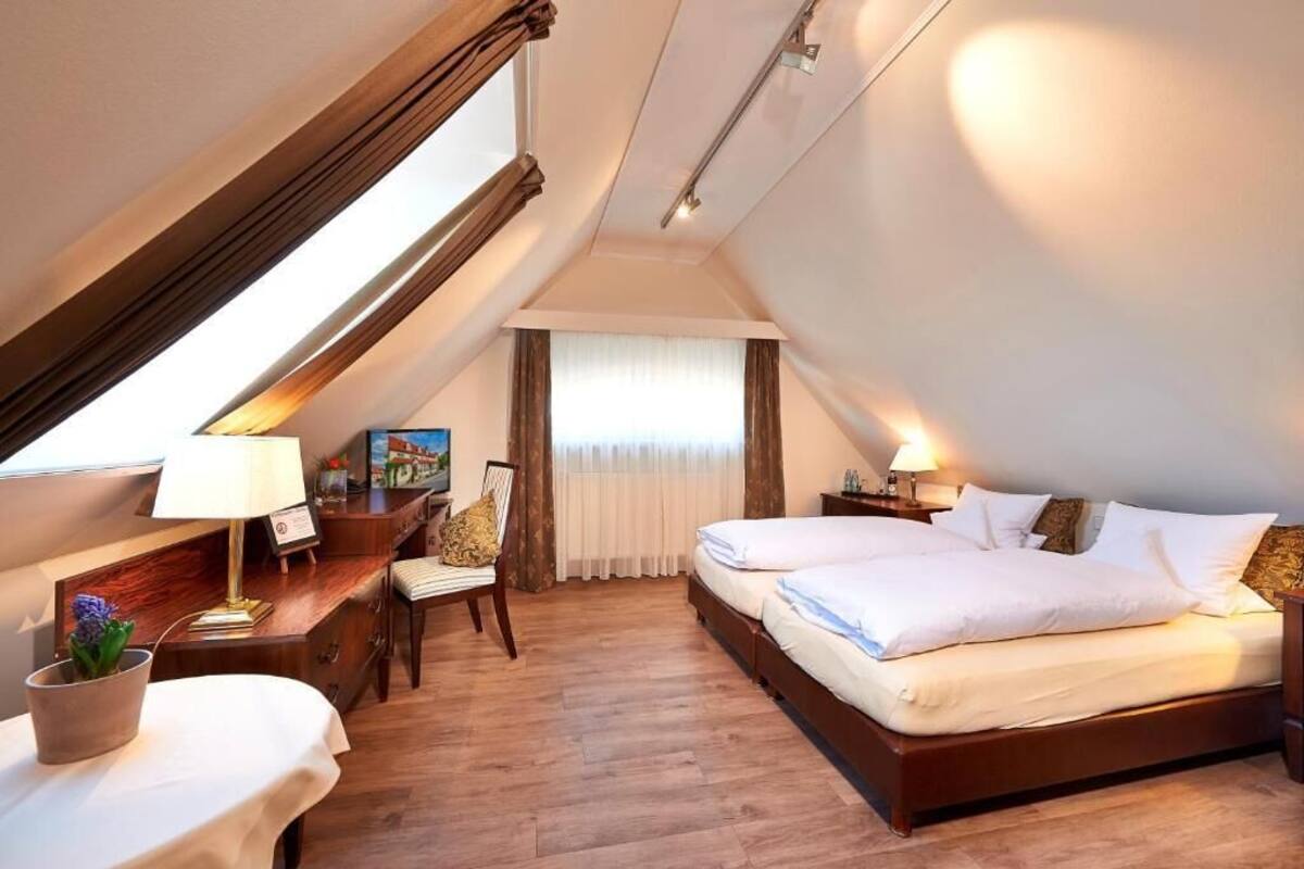 Habitación de hotel con una cama grande, un escritorio con lámpara, una silla, una mesita con una planta y una ventana con cortinas.