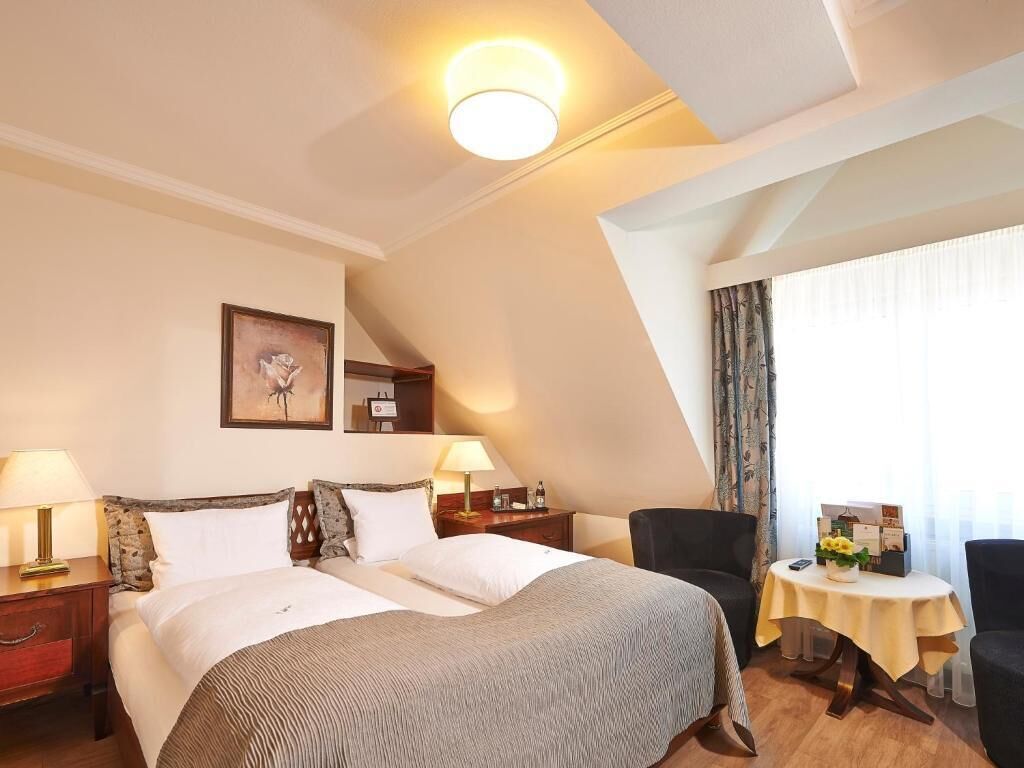 A hotel room with a bed, two bedside tables, a chair, a small round table, and a window with curtains.