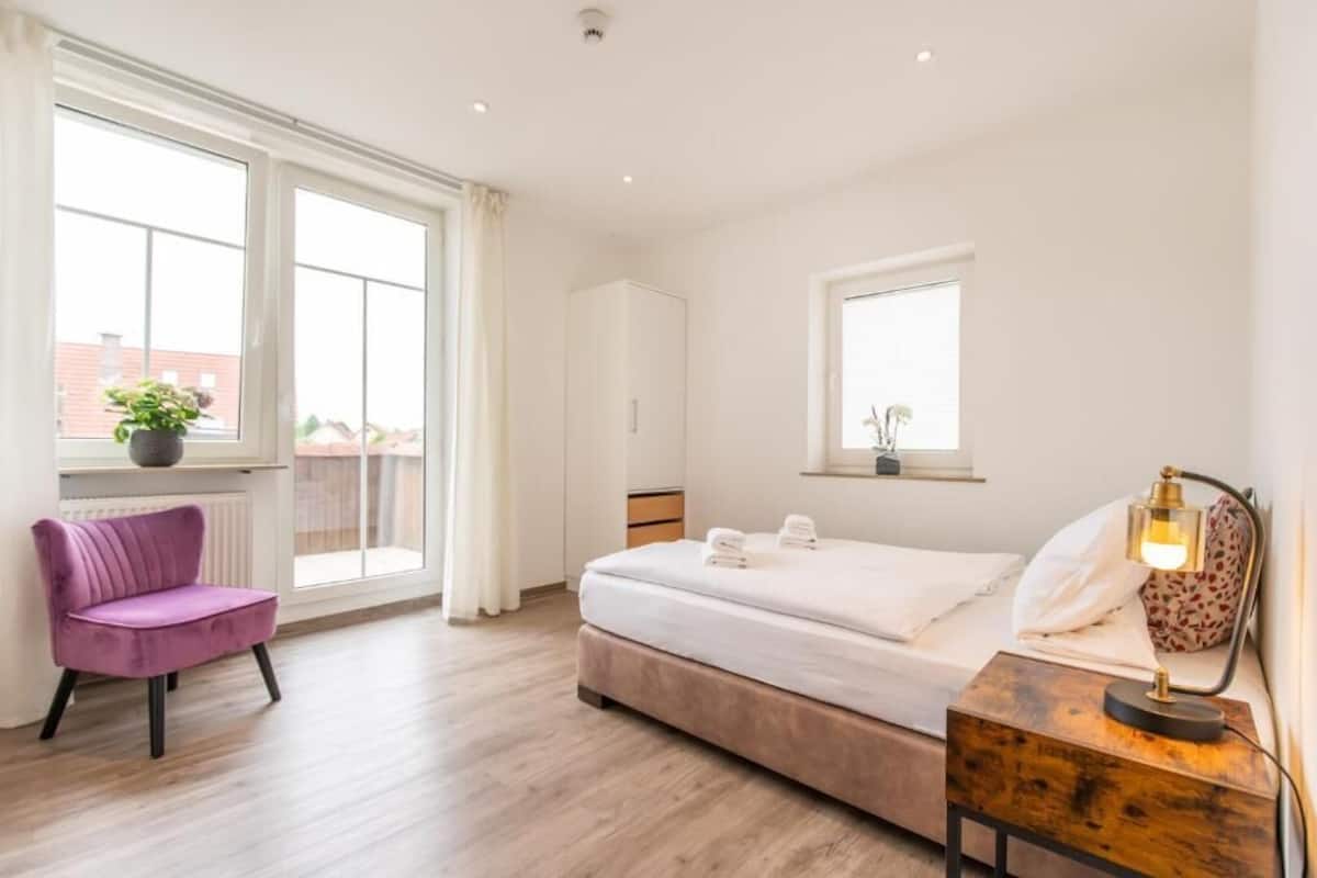Un dormitorio con cama, una silla, mesita de noche con lámpara, ventana y radiador.