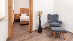 Habitación de hotel con piso de madera, una cama con ropa blanca, un sillón gris, un alto jarrón negro con plantas secas y un marco de puerta de madera.