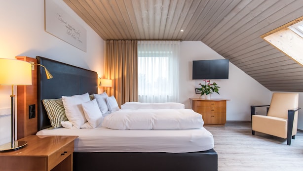 Un dormitorio moderno con una cama, mesitas de noche, un televisor, una silla y un ventanal con cortinas.