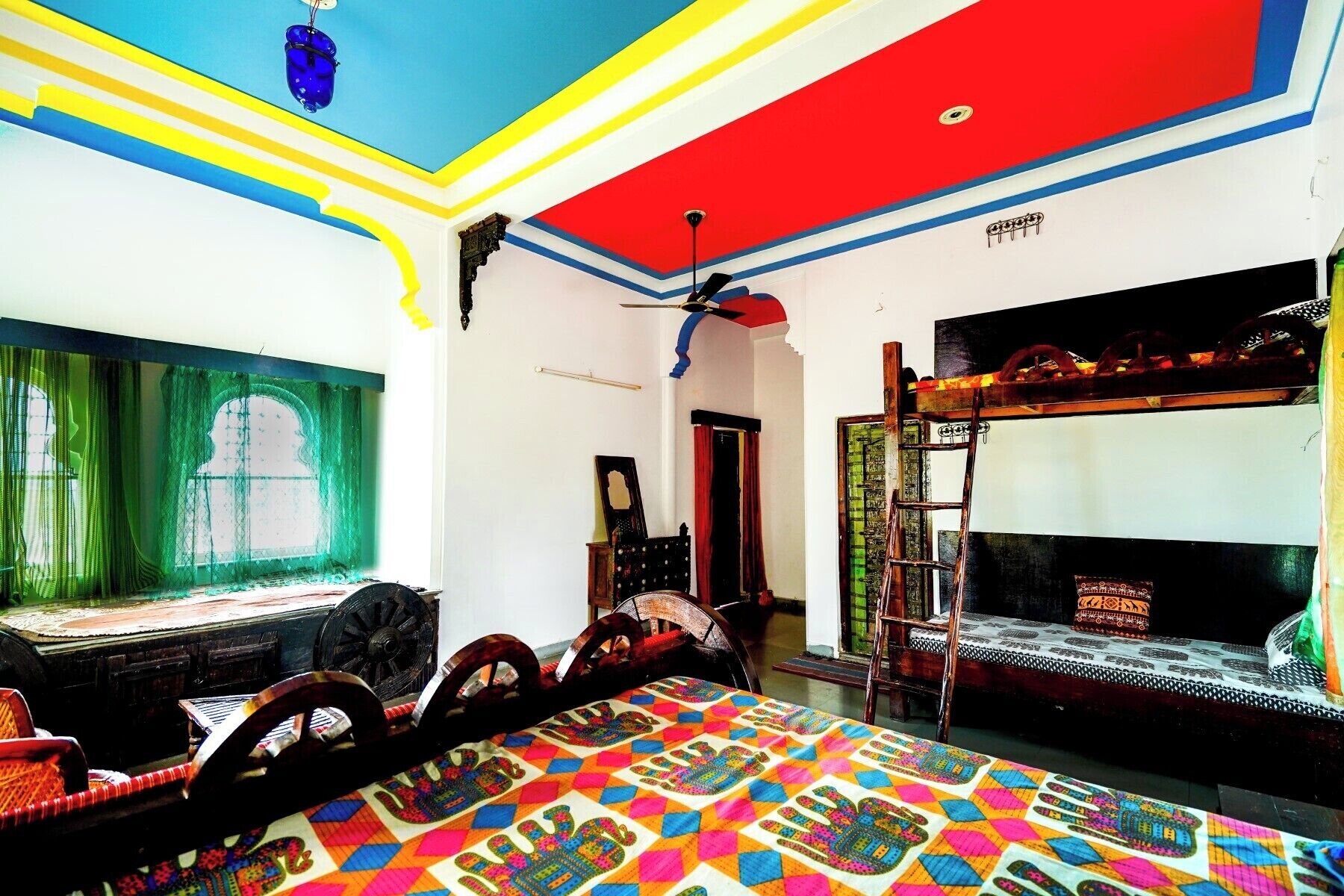 Una habitación con una cama litera, ventilador de techo, ventana con cortinas verdes y una colcha colorida en el suelo.