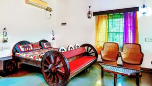 Een kamer met een bed, twee rieten stoelen, een kleine tafel, een plafondventilator en een raam met kleurrijke gordijnen.
