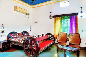 Une pièce comprenant un lit, deux fauteuils en osier, une petite table, un ventilateur de plafond et une fenêtre agrémentée de rideaux colorés.