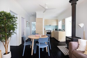 A modern kitchen with a central island, a dining area with blue chairs, a sofa, and a large potted plant. - Sunnyside - Forest path to beach (Bawley Point)