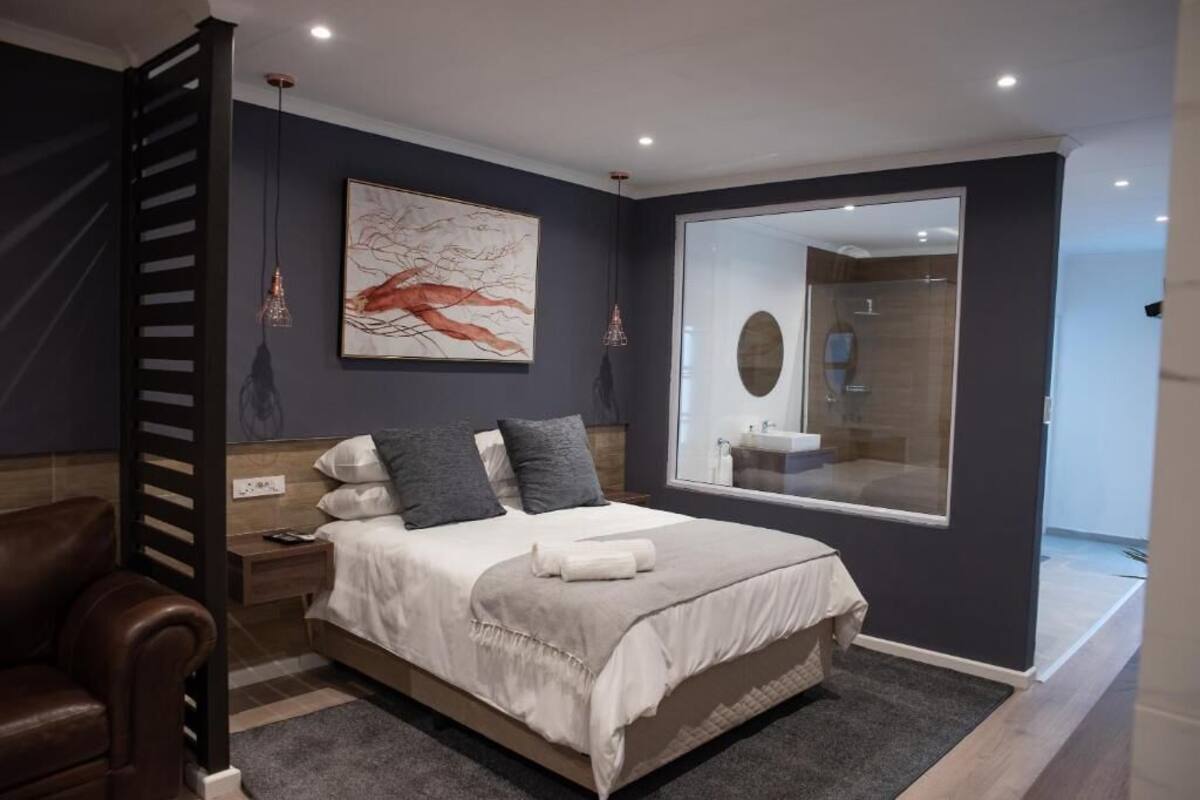 A modern bedroom with a large bed, a nightstand, a leather chair, a wall-mounted artwork, and a bathroom mirror.
