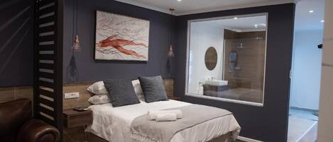 A modern bedroom with a large bed, a nightstand, a leather chair, a wall-mounted artwork, and a bathroom mirror.