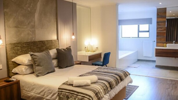 A modern bedroom with a large bed, bedside lamps, a desk, and a view of a bathroom. - The Royal Bryanston (Sandton)