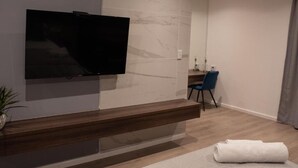 A modern room with a flat-screen TV mounted on the wall, a wooden bench, a blue chair, a small table with a mirror, and a rolled-up towel on the floor. - The Royal Bryanston (Sandton)
