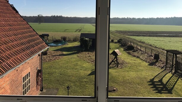 Blick aus einem Fenster auf ein grünes Feld, ein Backsteinhaus und einen klaren Himmel.
