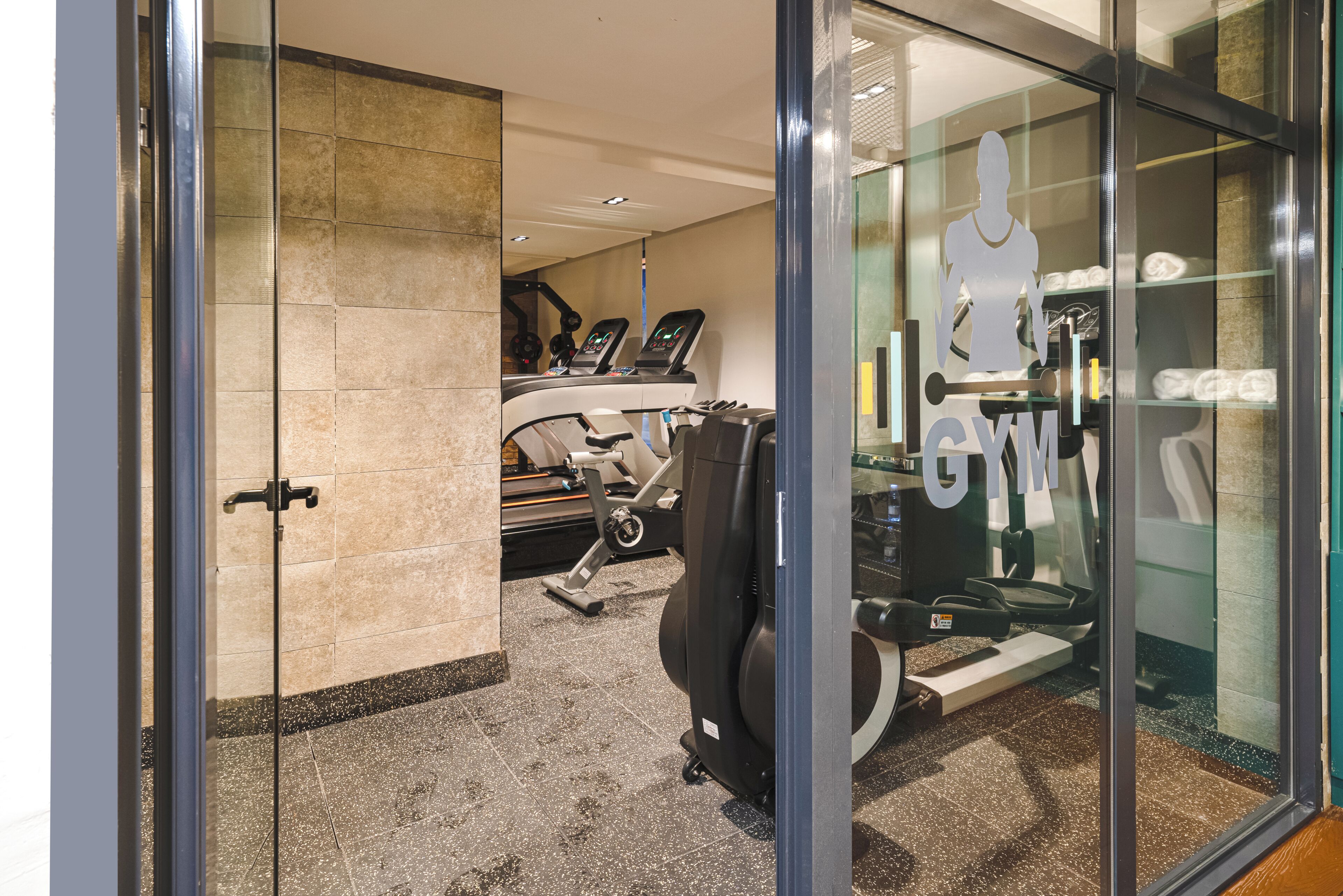 Un gimnasio con equipo de ejercicio, una entrada con puerta de vidrio y suelo de baldosas.