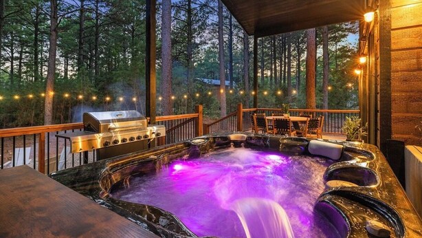 Un jacuzzi con caída de agua, rodeado por una terraza de madera y barandilla, con una parrilla y zona de estar a la vista.