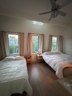 Une chambre avec deux lits, un ventilateur de plafond et de grandes fenêtres avec des rideaux.