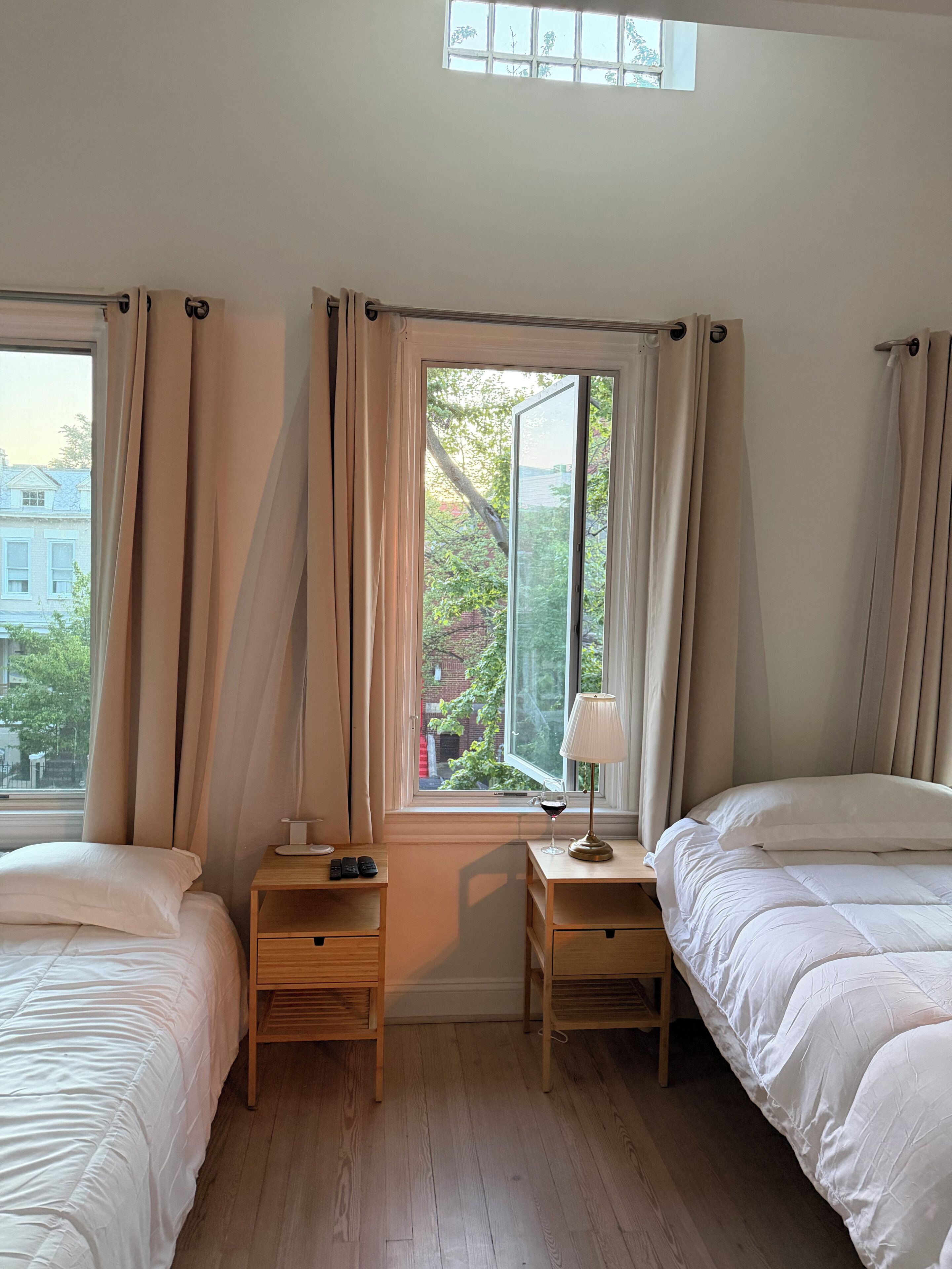 A bedroom with two beds, a window with curtains, a bedside table with a lamp, and a view of the outside.