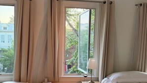 Ein Schlafzimmer mit zwei Betten, einem Fenster mit Vorhängen, einem Nachttisch mit Lampe und Blick ins Freie.