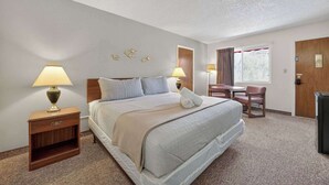 A hotel room with a bed, two bedside tables with lamps, a small table with a chair, a window with curtains, and a door. - Eagle Motel Suite 225 (Manitou Springs)