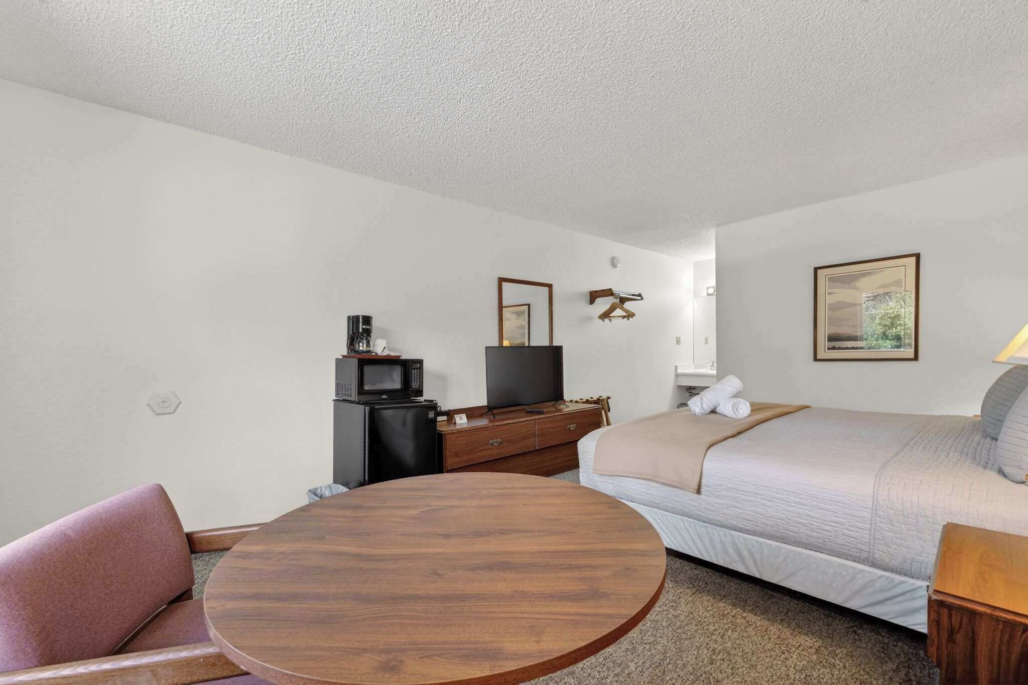 A hotel room with a round wooden table, a sofa bed, a TV, a dresser, and a painting on the wall.