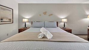 Ein ordentlich bezogenes Bett mit gefaltetem Handtuch, zwei Nachttischlampen und einem gerahmten Bild an der Wand.