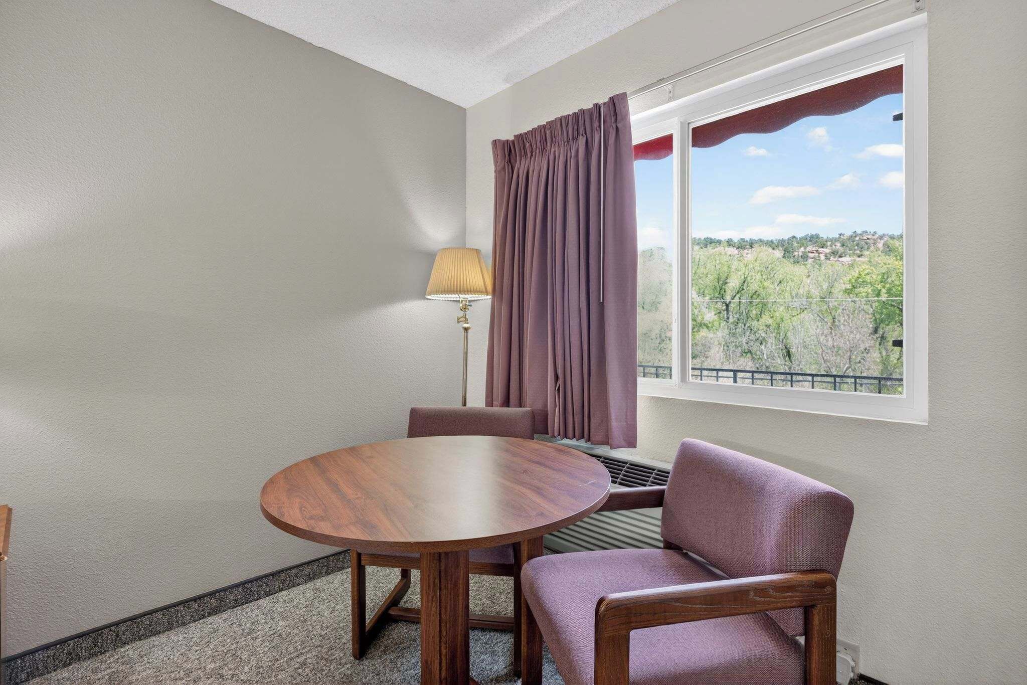 A room with a round wooden table, two chairs, a window with curtains, and a lamp.