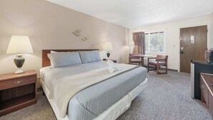 A hotel room with a bed, two bedside lamps, a desk with a chair, a window with curtains, and a door. - Eagle Motel Suite 226 (Manitou Springs)