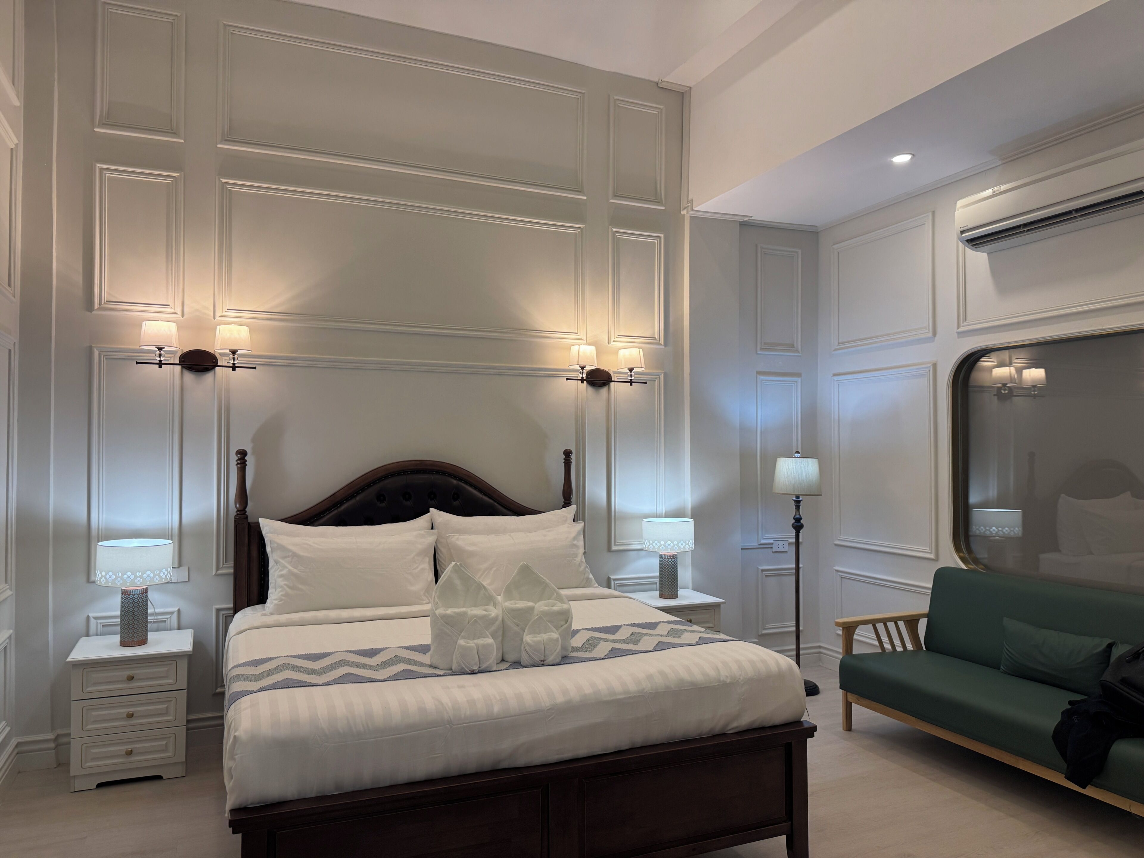 A neatly arranged bedroom with a bed, bedside tables, a nightstand, a green sofa, and an air conditioning unit.