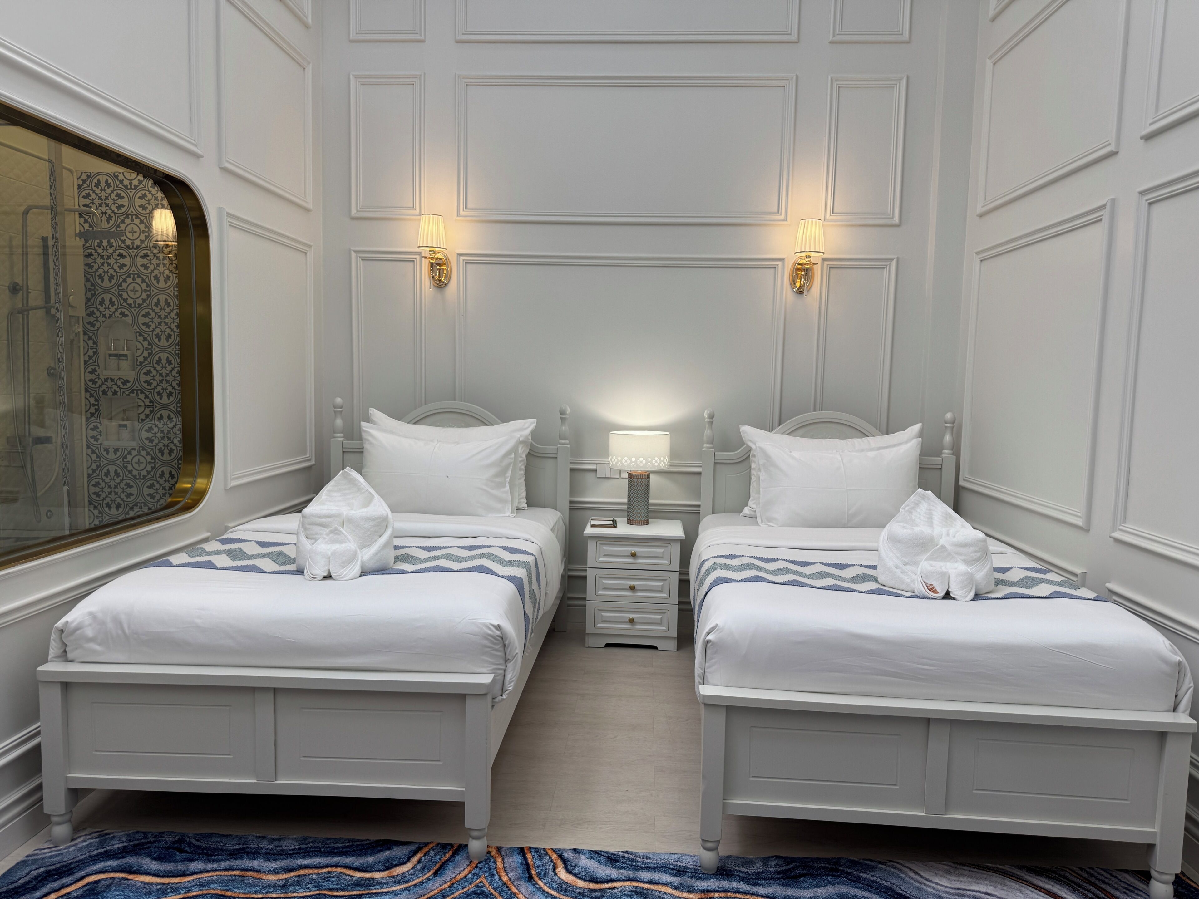 Habitación de hotel con dos camas, una mesita de noche con lámpara, un ventanal con vistas y una alfombra estampada.