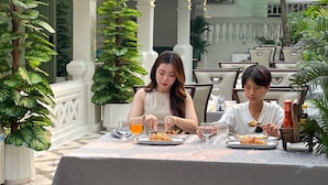 Two people dining at an outdoor table with a patterned tablecloth, surrounded by potted plants and white columns.