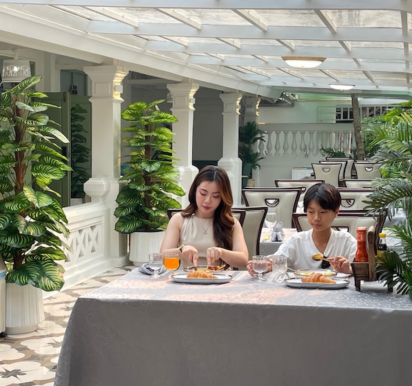 Dos personas cenando en una mesa al aire libre con mantel estampado, rodeadas de macetas con plantas y columnas blancas.