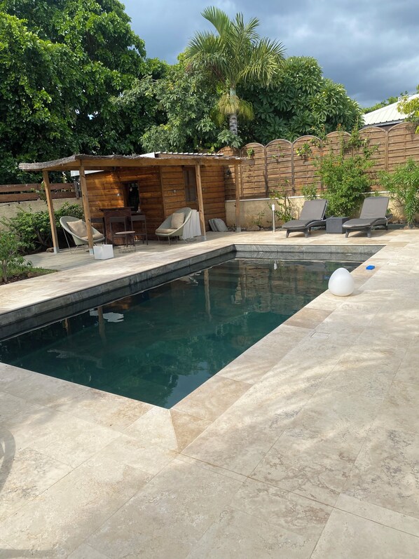 Una piscina rectangular con una terraza de madera, sillas de descanso y una pequeña estructura de madera.