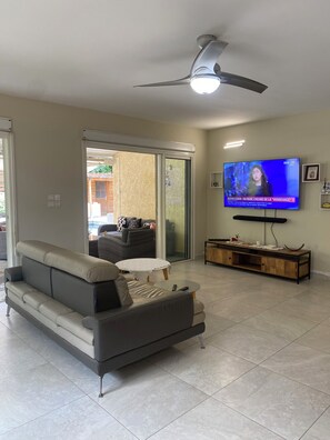 Una sala de estar con un sofá, una mesa de centro, un televisor, un ventilador de techo y un ventanal corredizo que da a un área exterior.