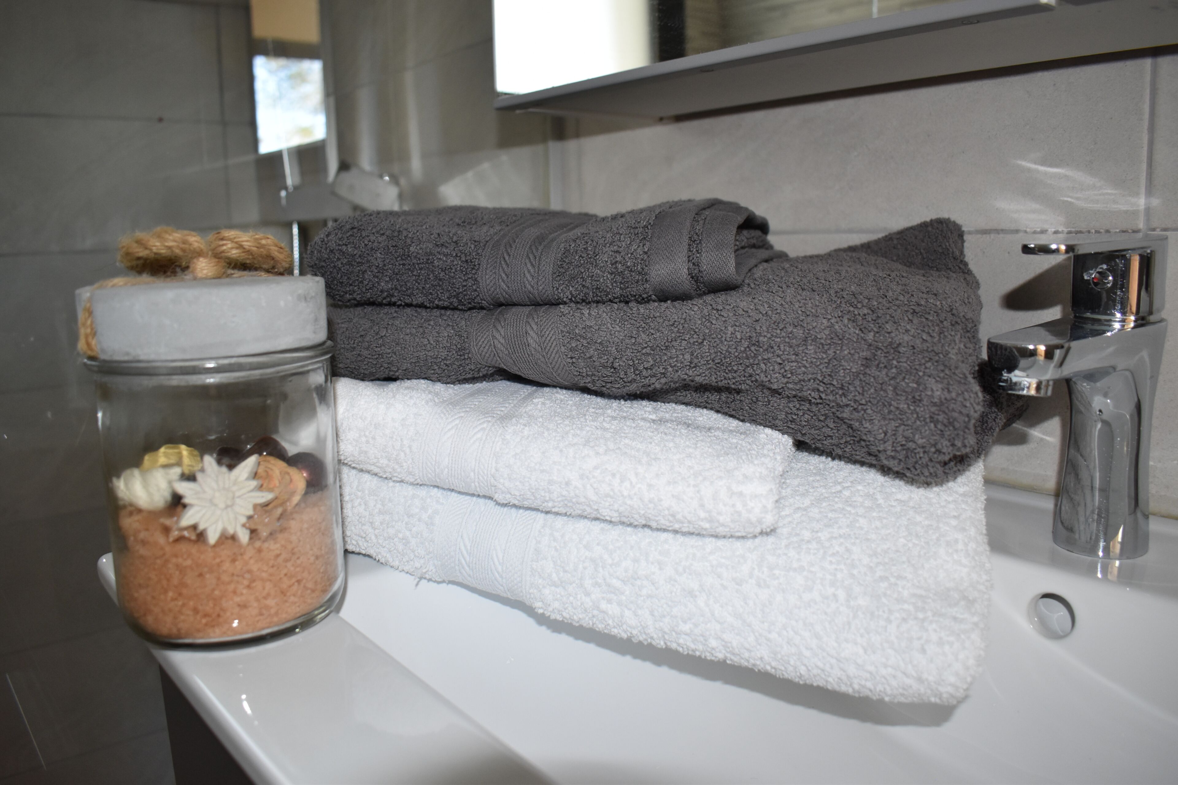 Un baño con un lavamanos blanco, un espejo y un tarro de vidrio con sales de baño y una flor blanca.