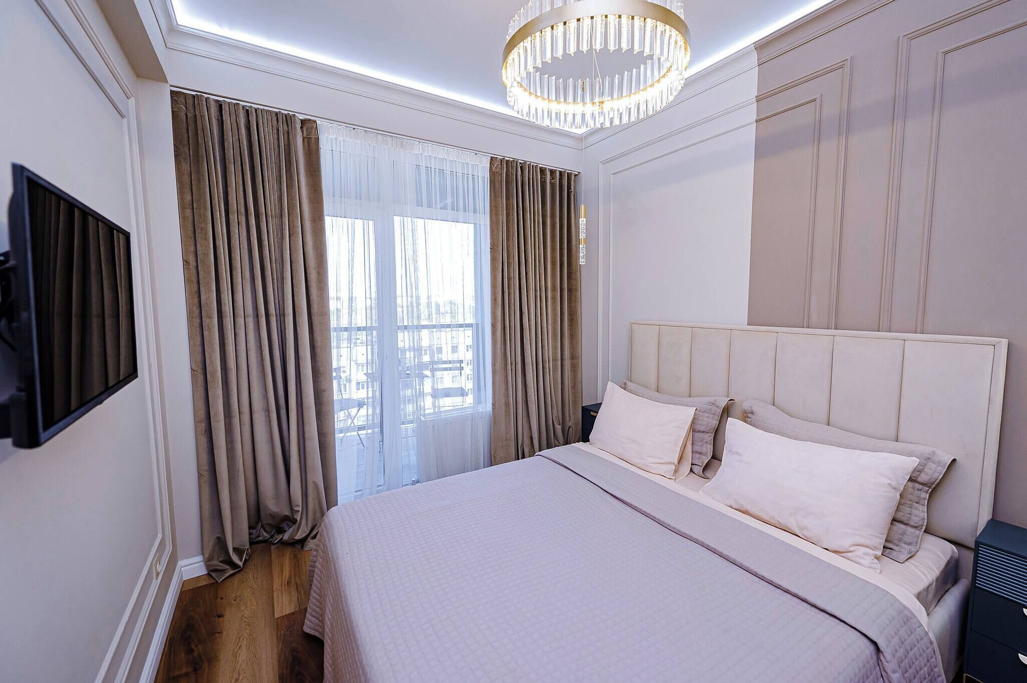 A hotel room with a large bed, a television mounted on the wall, and a chandelier.