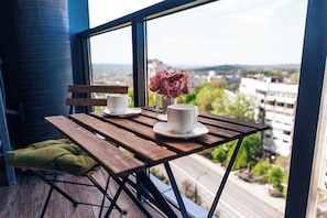 Un balcon avec une table en bois, deux tasses blanches, un verre et un vase rempli de fleurs.