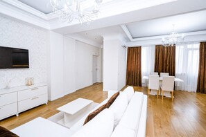 A modern living room with a white sofa, a dining table with chairs, a TV mounted on the wall, and a chandelier. - Luxurylakepark 2BD Apartment (Chișinău)