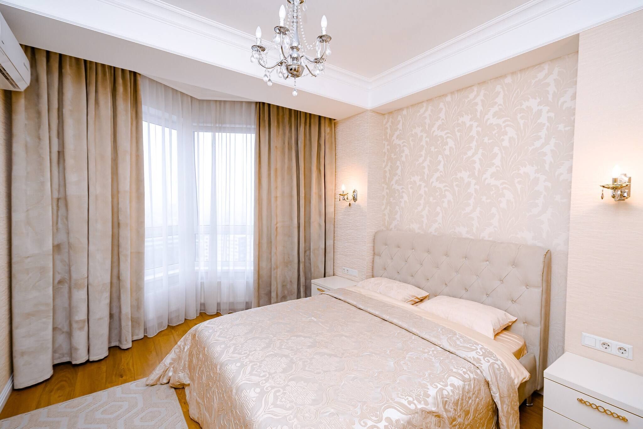 A bedroom with a large bed, a chandelier, curtains, and patterned wallpaper.