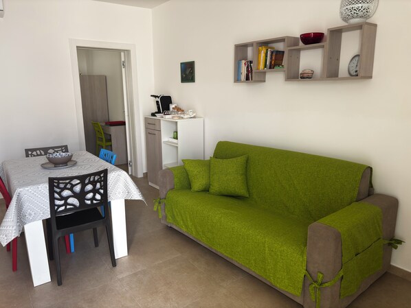 A living room with a green sofa, a dining table with chairs, and a kitchen area in the background.