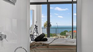 Un dormitorio con un ventanal grande con vistas al mar, una cama con manta a rayas y un trípode con una cámara.