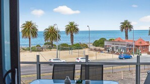 Vista desde una ventana sobre una playa con palmeras, una carretera con coches y un edificio con techo rojo.