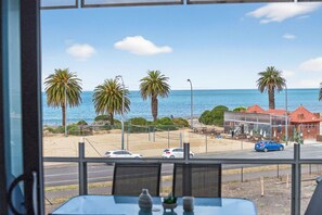 A view from a window overlooking a beach with palm trees, a road with cars, and a building with a red roof. - Beach House 305—Oceanfront Balcony Retreat (Victor Harbor)