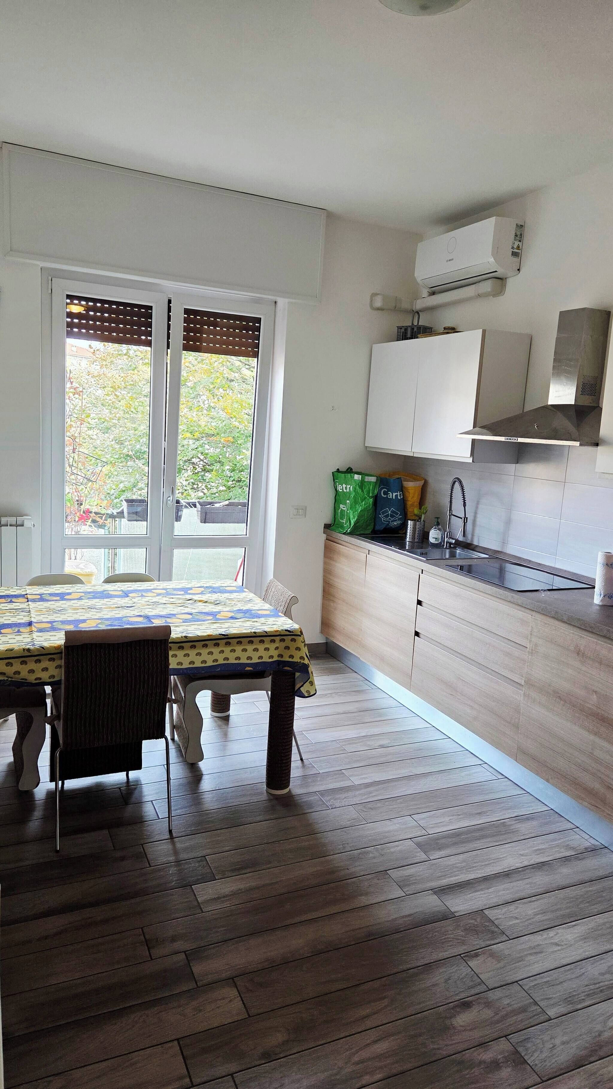 Una cocina con mesa, sillas, una ventana con persianas, un lavamanos, armarios y una bolsa de compras.