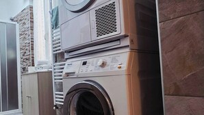 A compact, modern laundry room with a front-loading washer and dryer, a small countertop, and a tiled floor. - Charming Central Apartment Near Lake Maggiore in Arona (Arona)