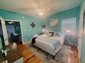 Une chambre à coucher comprenant un lit, un ventilateur de plafond, une fenêtre avec des stores et une porte donnant sur une autre pièce.