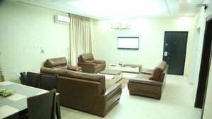 A living room with brown leather sofas, a dining table with chairs, a coffee table, and a TV mounted on the wall. - De Grandeur Tower Apartment (Ojodu)