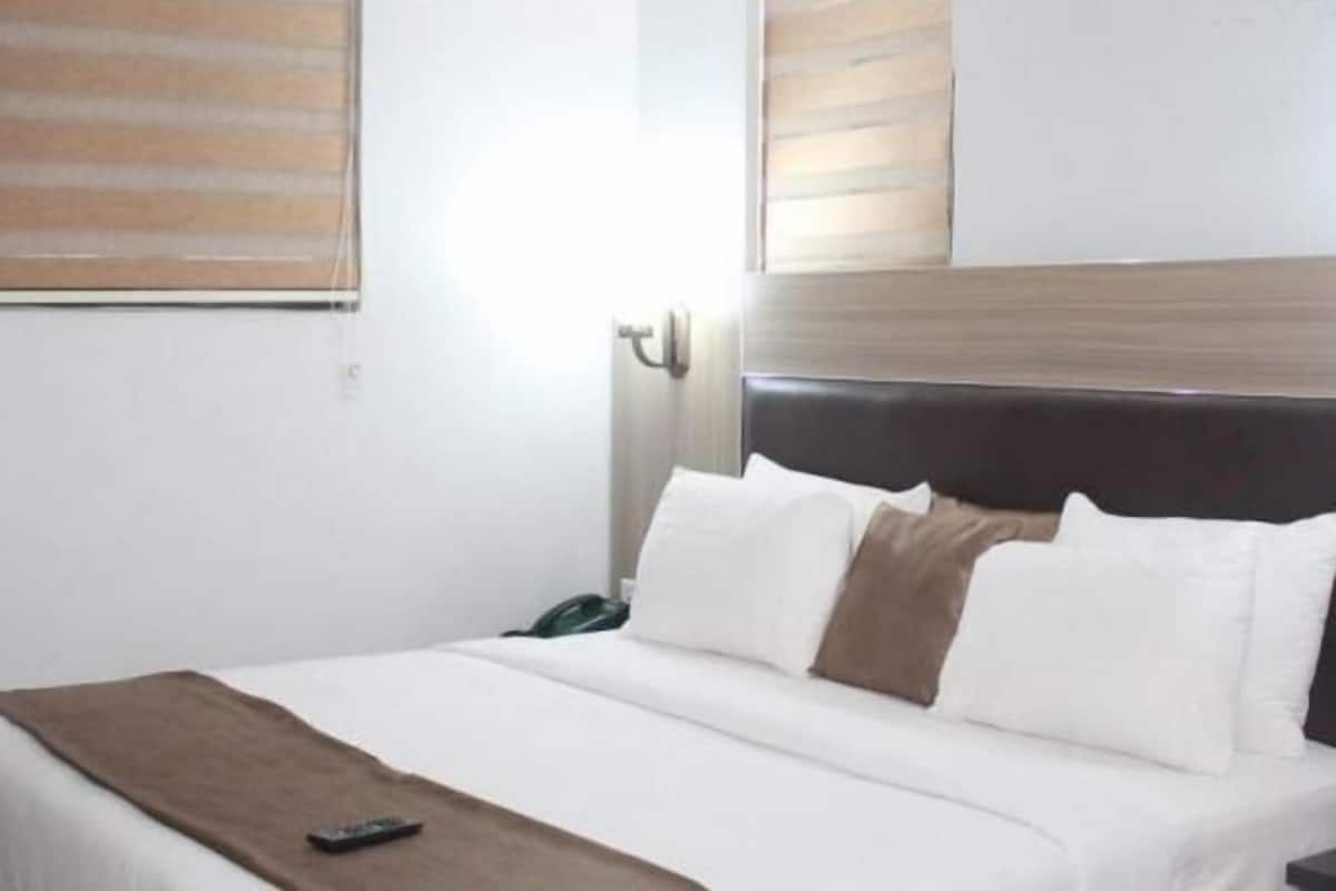 A hotel room with a bed, a bedside table, a remote control, and a window with blinds.