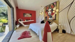 Ein modernes Schlafzimmer mit einem Bett, einer roten Akzentwand, einem großen Gemälde und Blick ins Grüne.