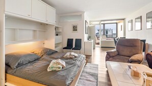 A bedroom with a bed, bedside tables, a chair, and a view of the living area.