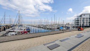 A marina with boats docked, a paved walkway, and a modern building with balconies.